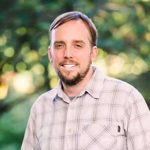 Angy Giles in a collared shirt smiling in front of a wooded background