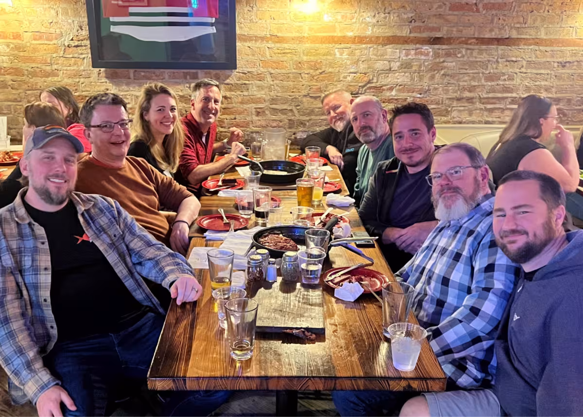 Group of people at a long table posing for a picture with pizza and beer on the table.